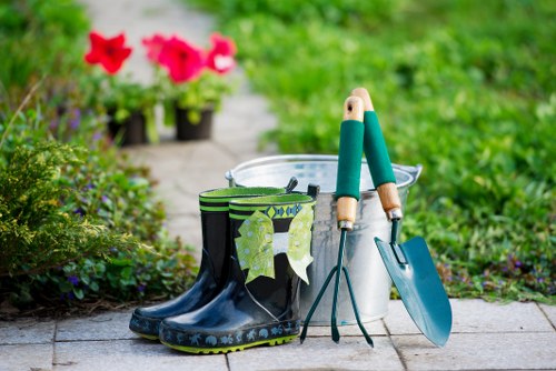 A pair of black rubber gardening boots with green leaf patterns, placed on a paved outdoor surface in a garden setting. The boots are slightly weathered and are situated next to various gardening tools, including a trowel with a teal handle, a hand fork, and a small spade, all leaning against a metal bucket. In the background, there are vibrant pink flowers in pots, along with lush green foliage and grass, indicating an actively maintained garden area. The scene suggests outdoor gardening activities typical of a landscaped backyard or front garden in a residential area near Edmonton, with natural lighting and a mild, bright weather condition. The environment shows a well-kept space, with a focus on planting and lawn care, aligning with professional gardening and landscaping services offered by Lawn Mowing Edmonton in the local area of the postcode for Edmonton in the UK.