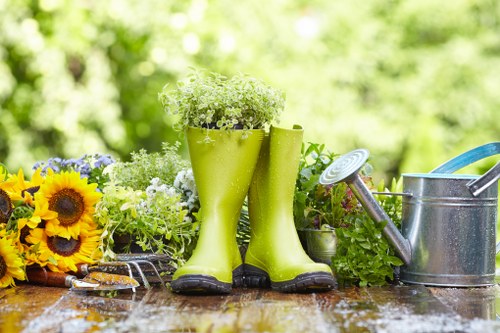 The image showcases a garden scene with a wooden outdoor table set against a lush, green backyard background. On the table, there are vibrant yellow sunflowers and various small white and purple flowering plants arranged in a container. Prominently positioned in the centre are a pair of bright green rubber boots filled with white flowering plants, serving as makeshift plant holders. To the right, there is a metal watering can with a curved spout, accompanied by additional gardening tools, including a small trowel and garden fork, indicating outdoor maintenance activity. The scene suggests a well-maintained garden area during daylight hours with natural sunlight highlighting the textures of the soil, plants, and garden accessories, emphasizing the connection to horticultural and landscaping services offered by Lawn Mowing Edmonton in the local area of Edmonton, UK postcode, and surrounding garden environment.